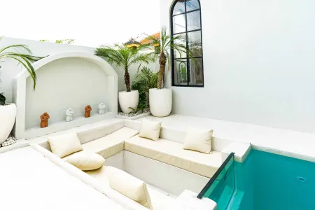 Modern white L-shaped outdoor sofa surrounding a turquoise infinity pool, with potted palms, terracotta statues, and arched tropical courtyard. - Bali Villas